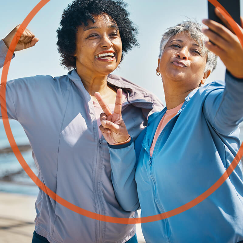 Zwei fröhliche Frauen mittleren alters in Outdoor-Sportkleidung. Sie posieren für ein Selfie. Die Frau mit dem Handy in der Hand macht ein Victory-Zeichen mit der freien Hand, die andere hat ihre Hand zu einer Faust geballt.