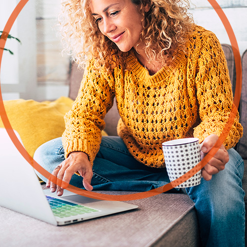 Lächelnde Frau mit offenen, lockigen Haaren, die mit einer großen Tasse in der Hand an einem Sofatisch sitzt und mit der anderen Hand ihren Laptop bedient.