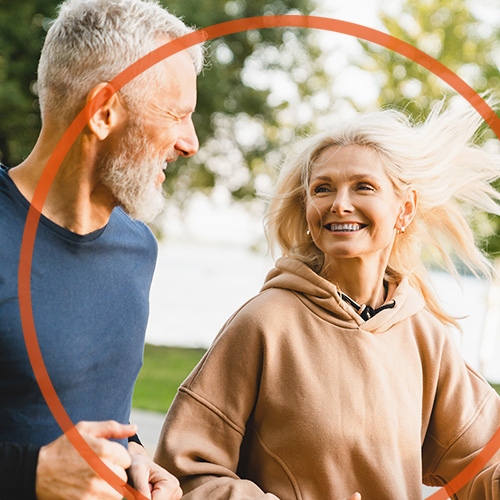 Frau beim entspannten Laufen draußen in der Natur. Sie ist im Fokus, neben ihr ein Mann im gleichen Alter. Sie schauen sich lächelnd an.